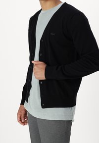 Černý kardigan vyrobený z pleteného materiálu, s výstřihem do V, knoflíkovým zapínáním a malým logem na hrudi. Nosený přes světle modrou košili.