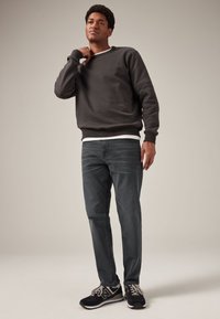 Jeune homme debout, portant un sweat-shirt noir, un jean gris foncé et des baskets noires, sur un fond neutre.