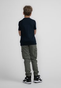 Marineblauwe T-shirt met korte mouwen, olijfgroene cargobroek met zijzakken, en zwarte hoge sneakers; krullend haar zichtbaar van achteren.