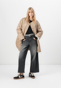 Mladá žena stojící v béžové oversized prošívané bundě, černém tričku, vybledlých širokých džínách a černo-bílých teniskách na bílém pozadí.