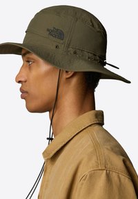 Zelený klobouk s širokým krempou a nastavovací šňůrkou na bradě, s logem The North Face. Nosený s béžovou košilí, která má texturovanou tkaninu.