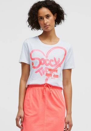 Frau mit lockigem Haar, die ein weißes T-Shirt mit pinkem Grafiktext und einen korallfarbenen Rock mit Kordelzug vor einem schlichten Hintergrund trägt.