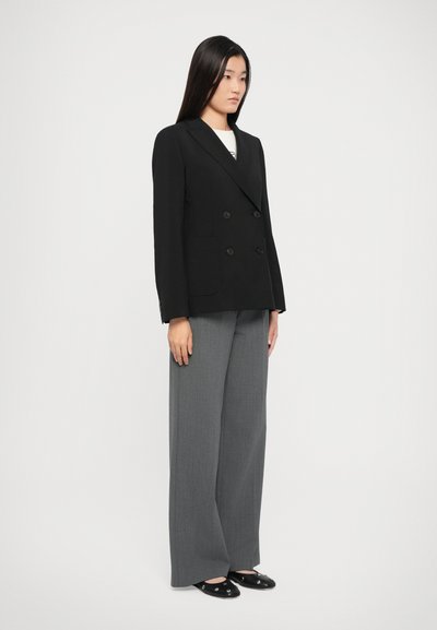 Schwarzes, doppelt-knöpfbares Blazer mit zwei vorderen Taschen, kombiniert mit grauen, weiten Hosen und schwarzen Slippern, vor einem weißen Hintergrund stehend.
