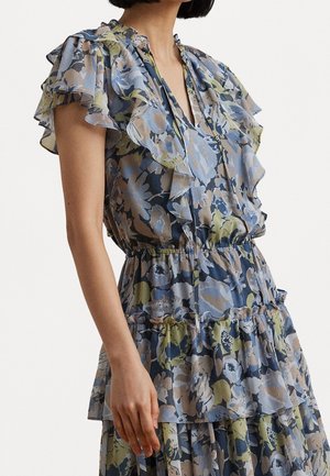 Kobieta w bezrękawowej, falbaniastej sukience w kwiaty w odcieniach niebieskiego, zielonego i beżowego, z warstwową tkaniną i talią podkreśloną paskiem.