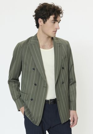 Olivengrøn dobbeltradet blazer med hvide striber, spidse revers, to forlommer og sorte knapper. Båret over en cremefarvet skjorte.