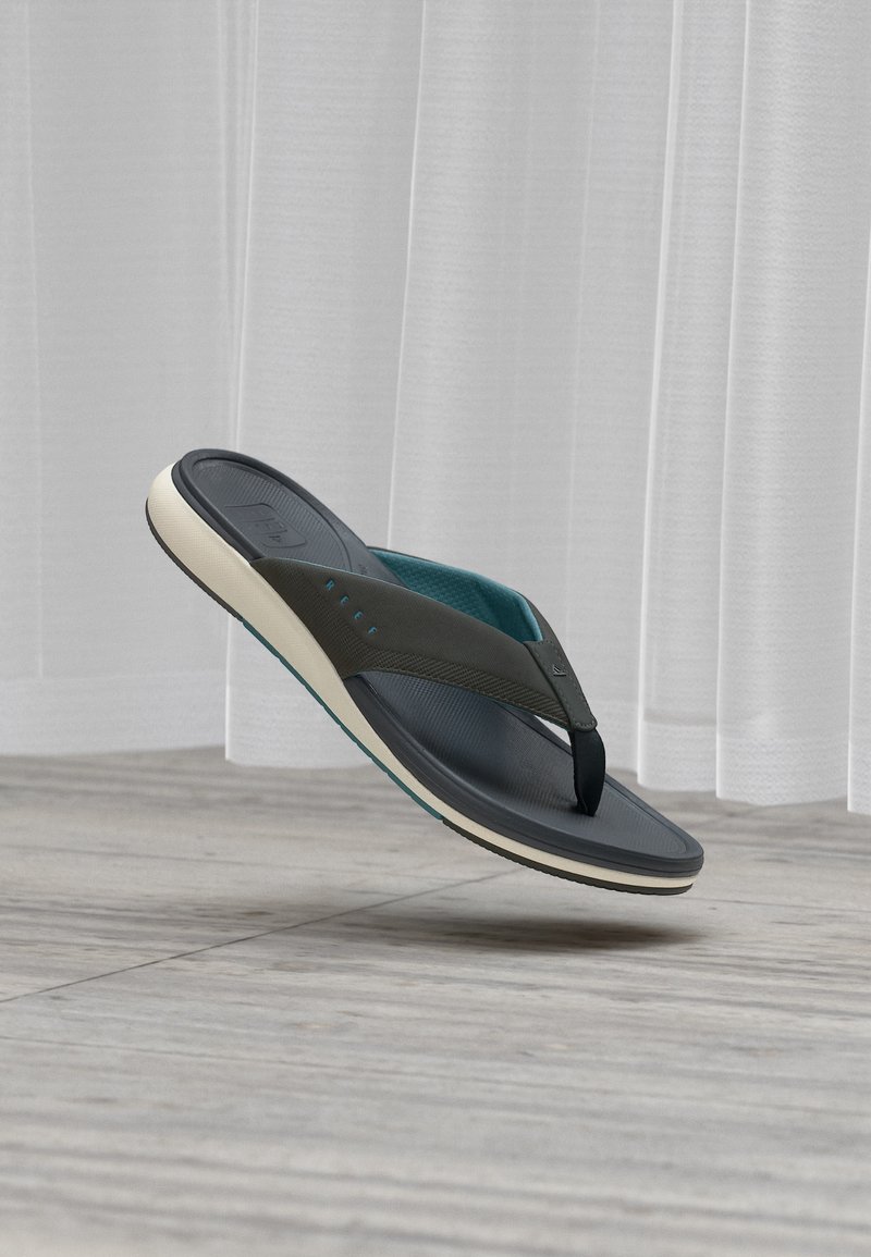 Μονό μαύρο και μπλε σαγιονάρι με λευκή σόλα που αιωρείται πάνω από γκρι ξύλινο πάτωμα με λευκές διαφανείς κουρτίνες στο φόντο.