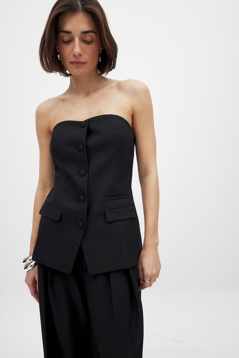 Musta olkaimeton paita, joka on valmistettu sileästä kankaasta. Siinä on kuusi etunappia ja kaksi sivutaskua, sekä muotoiltu malli ja jämäkkä siluetti.