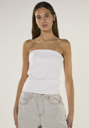 Młoda kobieta o długich brązowych włosach, ubrana w biały top bez ramiączek i jasnoszare jeansy z wysokim stanem, na jednolitym tle.