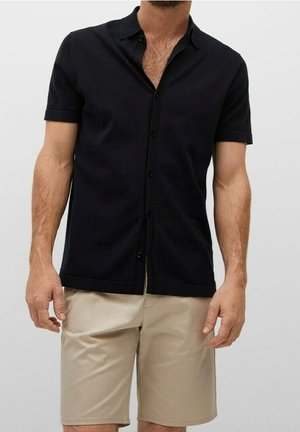 Mann trägt kurzärmliges schwarzes Hemd mit Knöpfen und beige knielange Shorts vor einfarbigem Hintergrund.