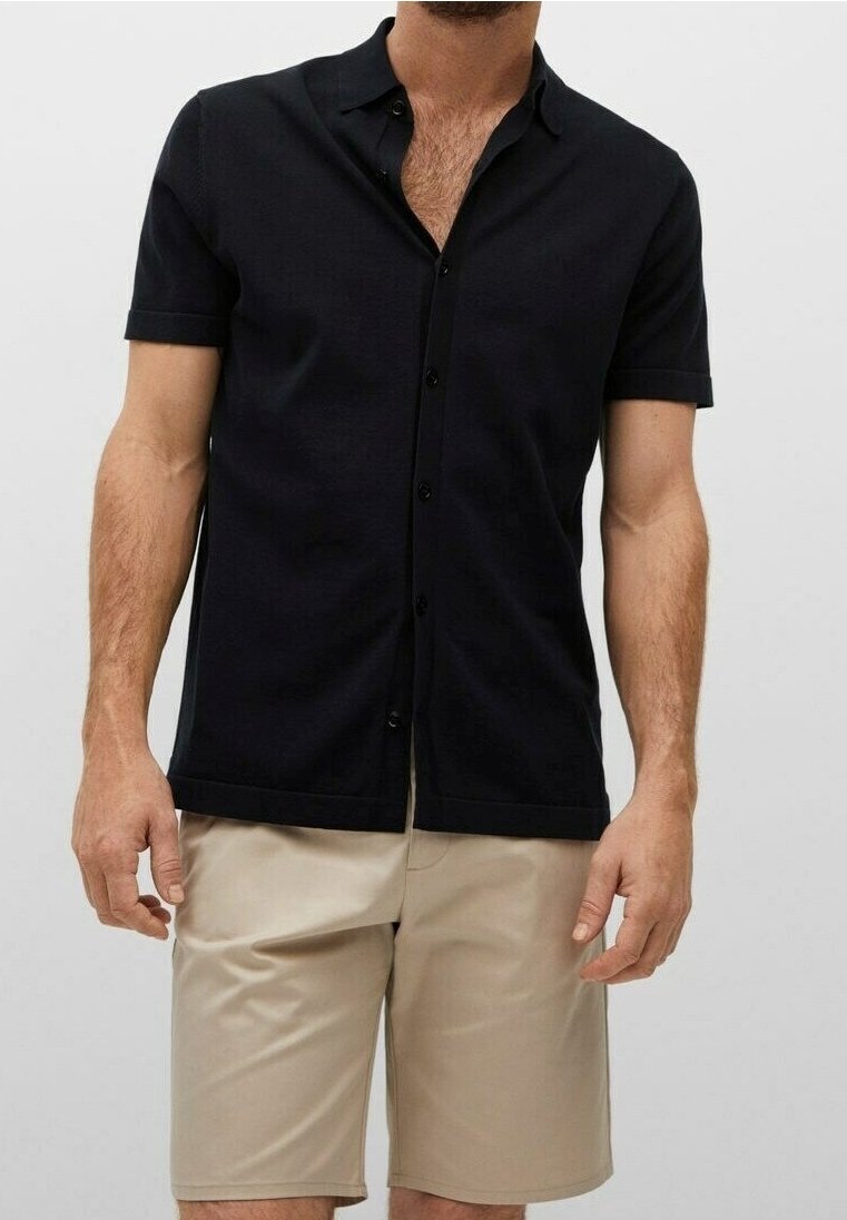 Homme portant une chemise noire à manches courtes boutonnée et un short beige jusqu’aux genoux sur un fond uni.