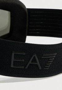 Közeli kép egy fekete szövetszíjról, rajta dombornyomott „EA7” felirattal és egy állítható elasztikus pánt részletével, fehér háttéren.