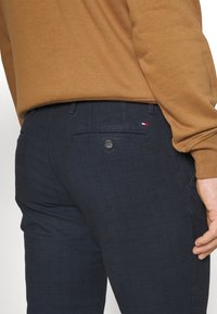 Marinblå rutiga byxor med en bakficka, knappdetalj och en liten röd, vit och blå logotypaccents i närheten av fickan.