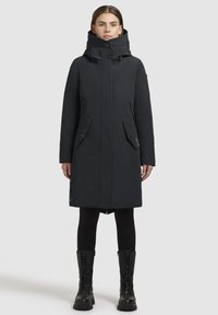 Manteau isolé noir avec un col haut, poches zippées et fermeture à boutons ; associé à des leggings noirs et des bottes en cuir à lacets arrivant au mollet.