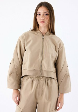 Beige cropped bomberjack met hoge kraag en ritssluiting, met een textuurstof en oversized mouwen. Gecombineerd met broek met elastische taille.