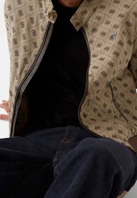 Primo piano di una persona che indossa una giacca beige a motivo con loghi marroni, aperta con zip sopra una maglietta nera e jeans blu scuro.