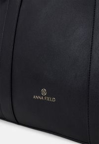 Schwarze Lederhandtasche mit glatter Textur, ausgestattet mit goldfarbenem "ANNA FIELD"-Logo und dezenten Nahtdetails an den Seiten.