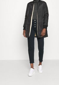 Svart vadderad lång jacka med krämfärgad foder och ribbade muddar, ihop med mörka jeans och vita sneakers, med knapprad.