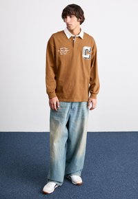 Camisa de polo de manga larga marrón con cuello blanco, que presenta texto bordado y un parche con la letra "C", combinada con unos pantalones vaqueros holgados de color azul claro.