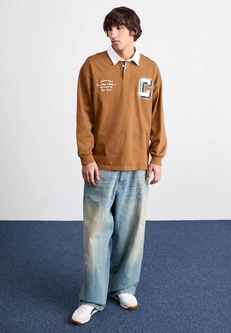 Braunes Langarm-Polohemd mit weißem Kragen, versehen mit gesticktem Text und einem Buchstaben "C"-Patch, kombiniert mit hellblauen weit geschnittenen Jeans.