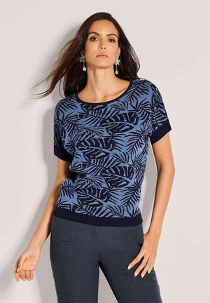 Frau trägt ein blau-schwarzes, gemustertes Kurzarmoberteil und dunkle Hosen, dazu baumelnde Blumenohrringe, posiert vor einer hellen Wand.