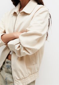 Chaqueta bomber beige con cremallera frontal, dobladillo acanalado y mangas fruncidas. Tejido liso y diseño minimalista con bolsillos laterales.