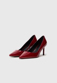 Raudoni lakuoti aukštakulniai batai su smailiu nosiu, elegantiška siluetu ir plonu stiletu. Juodas vidinis pamušalas.
