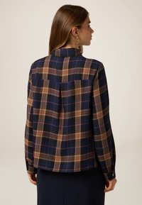 Camisa de cuadros en colores navy, marrón y crema. Presenta un cuello, puños con botones y un yugo trasero con un pliegue central. Textura de tela suave y tejida.