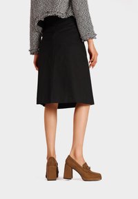 Scarpe a blocco di tacco in suede marrone con punta rotonda e lacci decorativi, abbinate a una gonna nera fino al ginocchio e a un maglione grigio texturizzato.