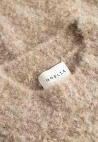 Smėlio spalvos vilnonė tekstūruota medžiaga su siuvine prisiūta balta drabužių etikete, kurioje juodais rašmenimis užrašytas prekinis ženklas „NOELLA“.