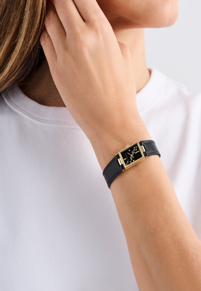 Fekete bőrszíjjal rendelkező óra arany téglalap alakú tokban, fekete számlappal, amely arany részleteket tartalmaz. A szíj letisztult és egyszerű.