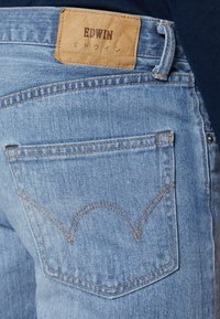 Gros plan de la poche arrière d'un jean en denim bleu avec une étiquette en cuir marron portant l'inscription Edwin et des caractères japonais.