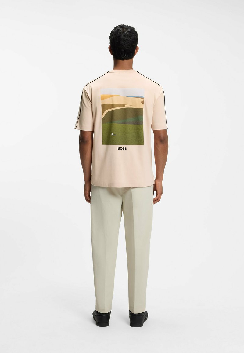 Homme de dos face à la caméra, portant une chemise beige avec un motif de parcours de golf et le texte "BOSS", un pantalon clair et des chaussures noires.