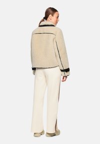 Beige shearling jacket with black accents, cropped design, and wide sleeves. Paired with cream trousers featuring side slits.