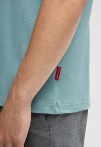 Polo en tissu texturé bleu clair, à manches courtes, avec une étiquette rouge "JOOP! JEANS" près de l'ourlet, associé à un pantalon gris.