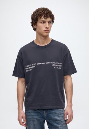 Jeune homme aux cheveux bouclés portant un t-shirt gris foncé avec un texte blanc et un jean bleu, regardant vers la gauche sur un fond uni.