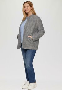 Graue Wollmischungjacke mit rundem Halsauschnitt, Fronttaschen und lässiger Passform. Getragen über einem blauen Strickpullover und kombiniert mit Jeans.