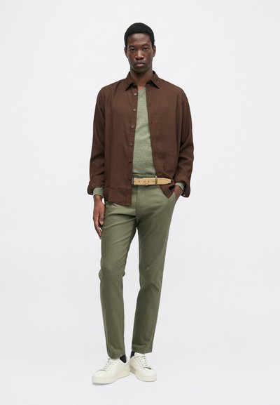 Jovem em pé, vestindo uma camisa marrom de botões sobre um suéter verde, calças olivas com cinto bege e tênis brancos, em fundo branco.