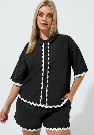 Kvinde med glat blondt hår iført sort skjorte med knapper og shortsæt med hvid bølget kant, poserer mod ensfarvet baggrund.