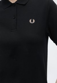 Polo noir avec une patte à deux boutons et un petit emblème de couronne de laurier blanche sur la poitrine gauche.