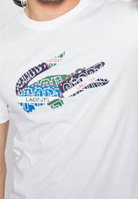 White cotton t-shirt featuring a multicolored graphic of a crocodile logo with text elements in blue, green, red, and black.