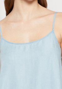 Světle modrý top bez rukávů vyrobený z jemné tkaniny, s tenkými nastavitelnými ramínky a kulatým výstřihem s jemným leskem.