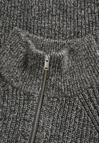 Gros plan sur un pull en maille gris et noir avec un motif côtelé et une fermeture éclair centrale en métal argenté partiellement fermée.