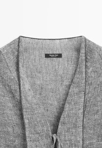 Grau texturierter Leinenjackenkragen mit sichtbarem schwarzem Etikett mit der Aufschrift "Massimo Dutti Made in China" auf weißem Hintergrund.