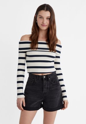 Olkapäiltä avonainen neule, jossa on tummansinisiä ja valkoisia vaakasuoria raitoja, ribbitekstuuria, yhdistetty korkeavyötäröisten mustien shortsien kanssa.