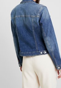 Chaqueta de denim en un lavado azul medio, con un corte clásico, cierres de botón en los puños y detalles bordados a lo largo de los hombros.