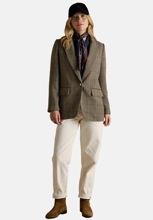 Brown plaid blazer with a single gold button, cream trousers, and brown ankle boots; accessorized with a dark cap and floral-patterned scarf.