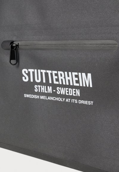 Borsa impermeabile grigia con una tasca frontale con zip e testo bianco in grassetto che recita "STUTTERHEIM" e "STHLM - SVEZIA." Materiale testurizzato con finitura opaca.