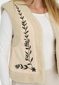 Beige fleeceväst med mjuk textur och öppen front, prydd med svarta broderade bladmönster på en beige panel.