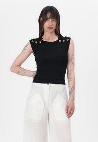 Black ribbed tank top with gold button accents on shoulders, paired with light-colored wide-leg pants featuring large front pockets.