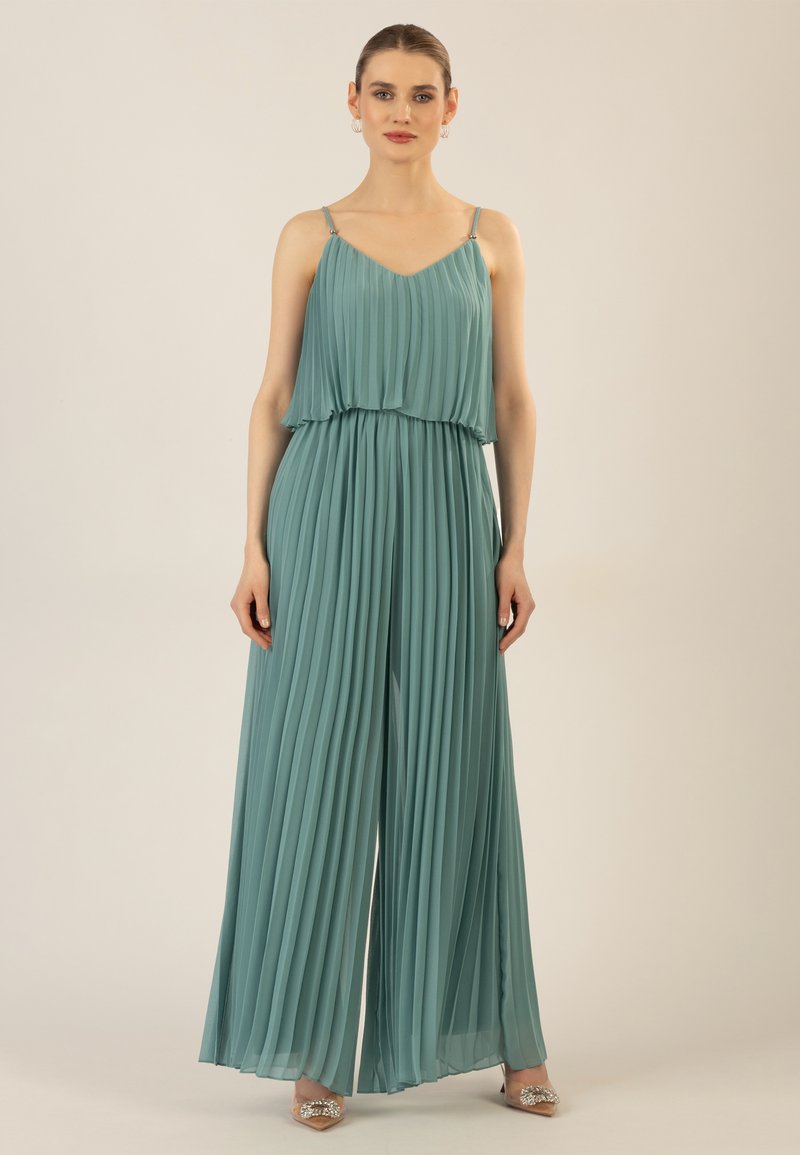 Een teal geplooide jumpsuit met een vloeiende ontwerp, dunne banden en een getailleerde taille, aangevuld met versierde nude hakken.